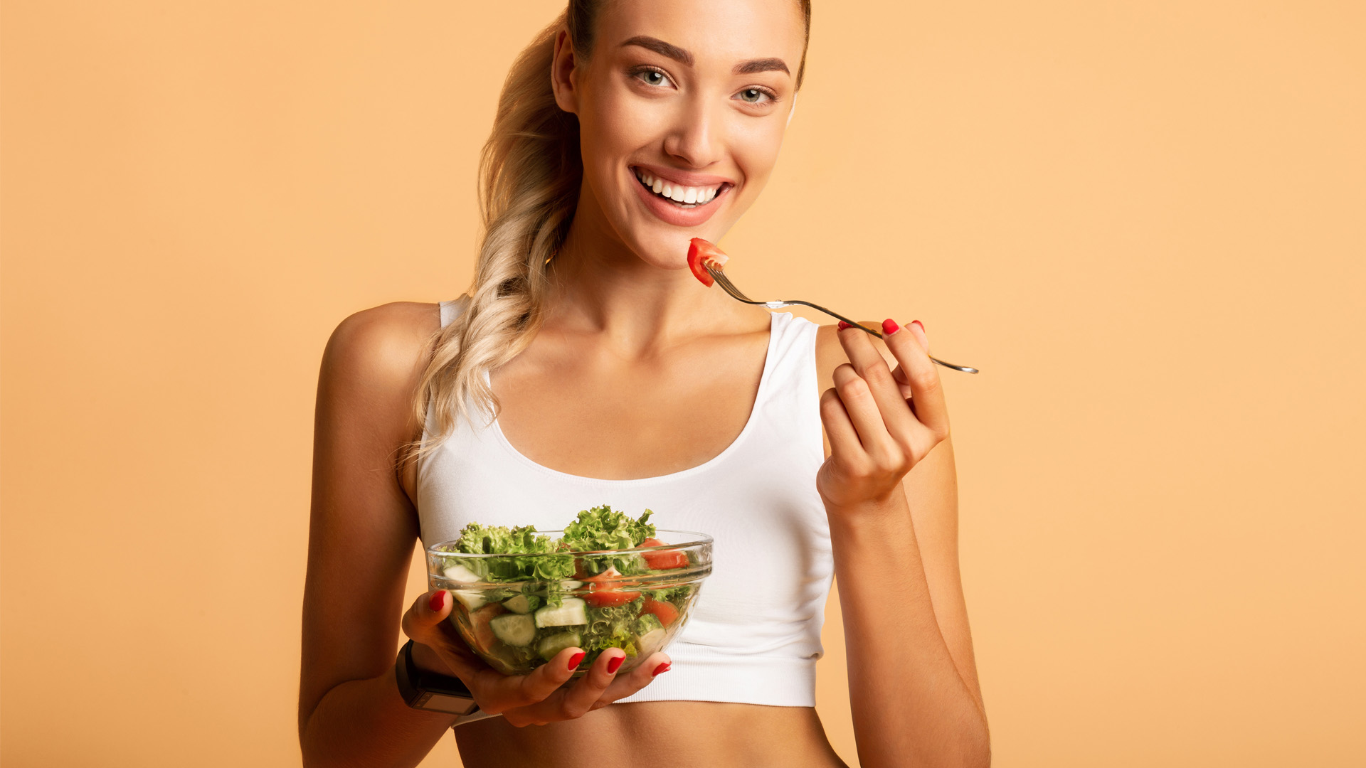 Donna sorridente mangia insalata fresca con pomodori e cetrioli, indossando top bianco su sfondo arancione.